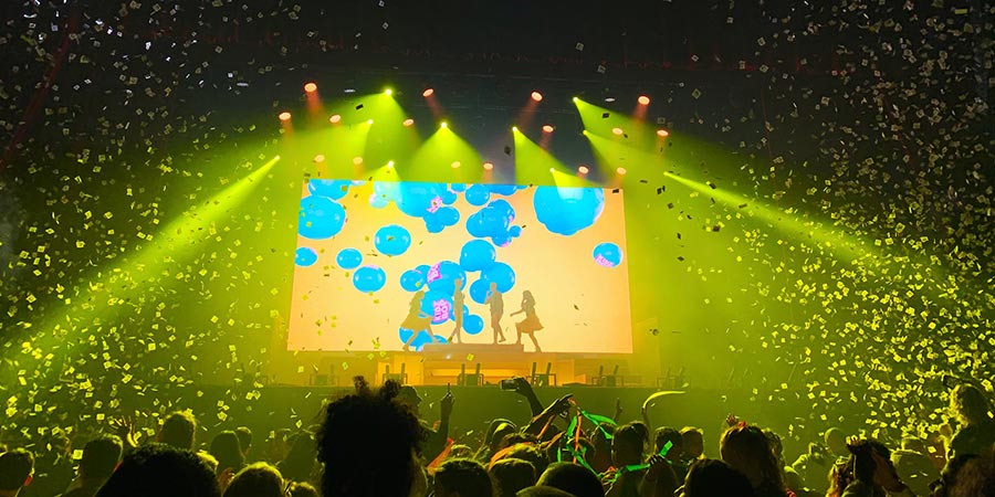 four people on stage in front of a crowd with yellow lighting and a big screen at the back
